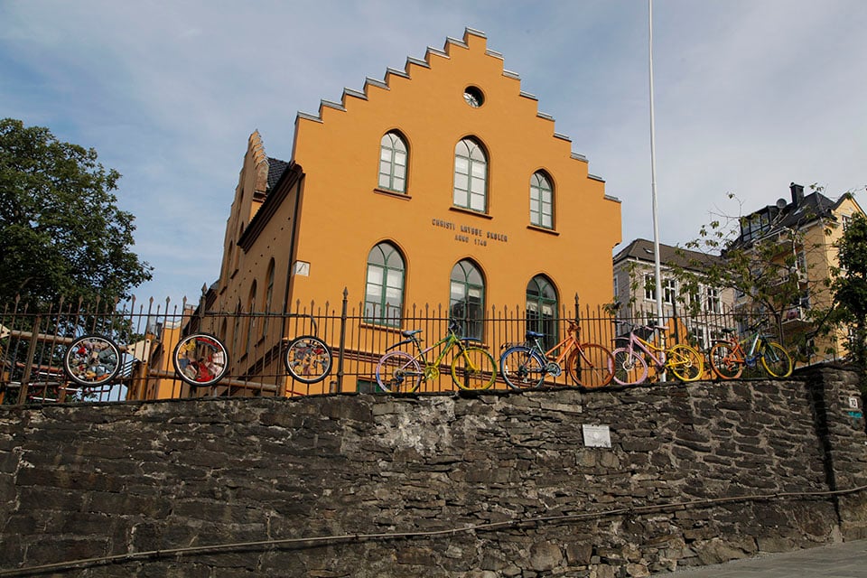 A bright orange-yellow building with a jagged, stepped roof stands behind a low stone wall. In front of the building, a row of colorful bicycles, painted yellow, blue, and pink, are lined up behind a black metal fence. The fence is decorated with several bicycle wheels.